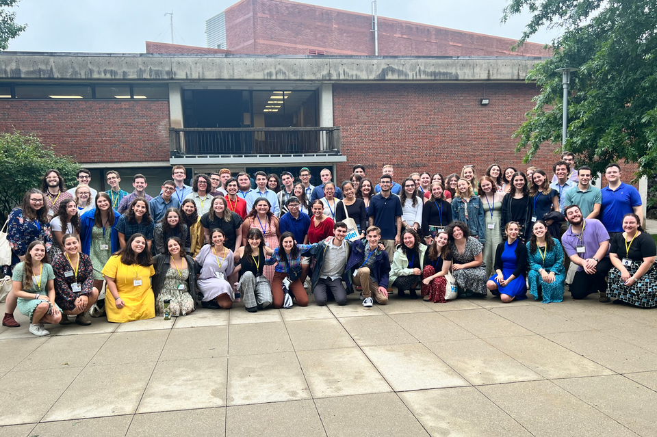 Very large group of people standing and kneeling outside a brick building smiling at the camera