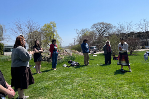 People stand in a loose circle outside on the grass with a rock in the background, with a native woman in a skirt with holding something