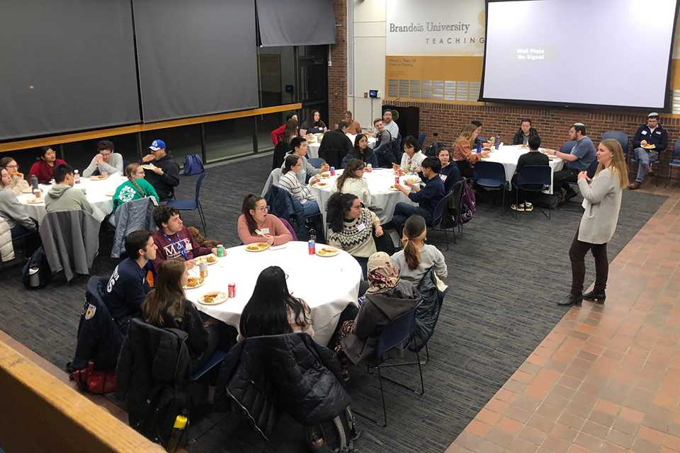 large room with multiple tables of students sitting around them, with a speaker in front and a large screen pulled down on one side