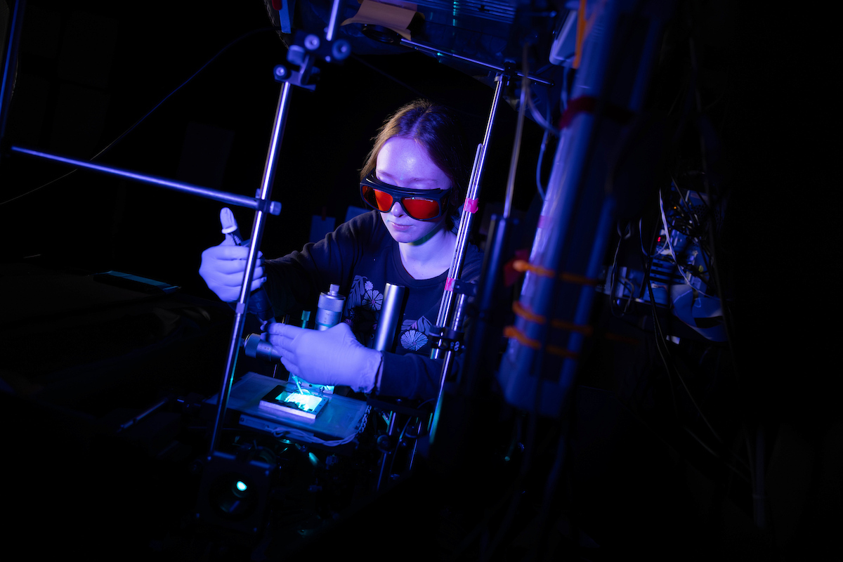 Fiona Stewart stands in front of a table covered in lenses and mirrors. She is wearing a pair of tinted safety glasses, and is adding microbes to a plate of glass using a pipette.