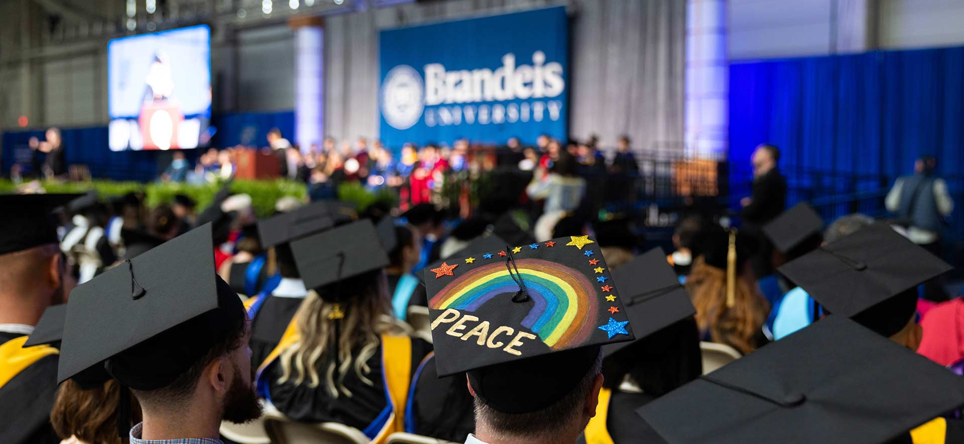 Close up of a graduation cap with a rainbow and the word "Peace" with the Commencement stage in the background