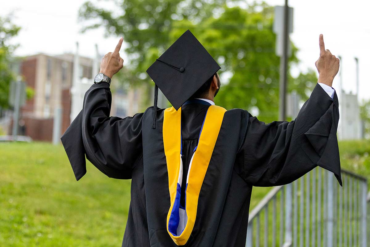 The back of a person in Commencement attire, holding up an index finger on each hadn