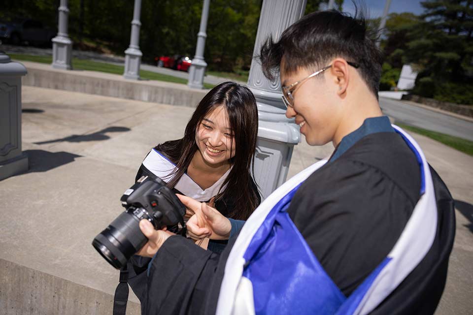 Graduates dressed in Commencement attire smile and look at photos on a camera.