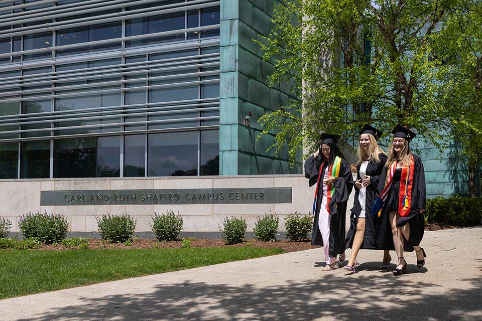 Three graduates dressed in Commencement attire walk together on the Brandeis campus.
