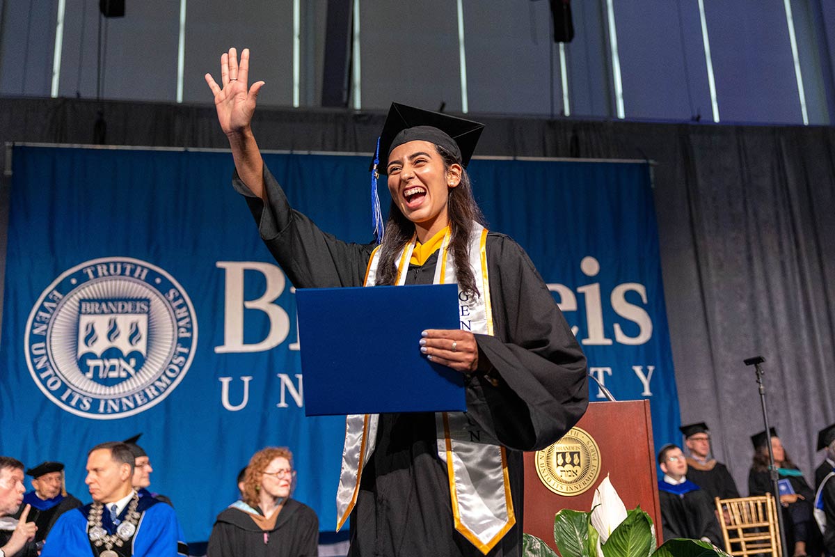 An excited graduate walks across the stage at Commencement waving and holding their diploma. 