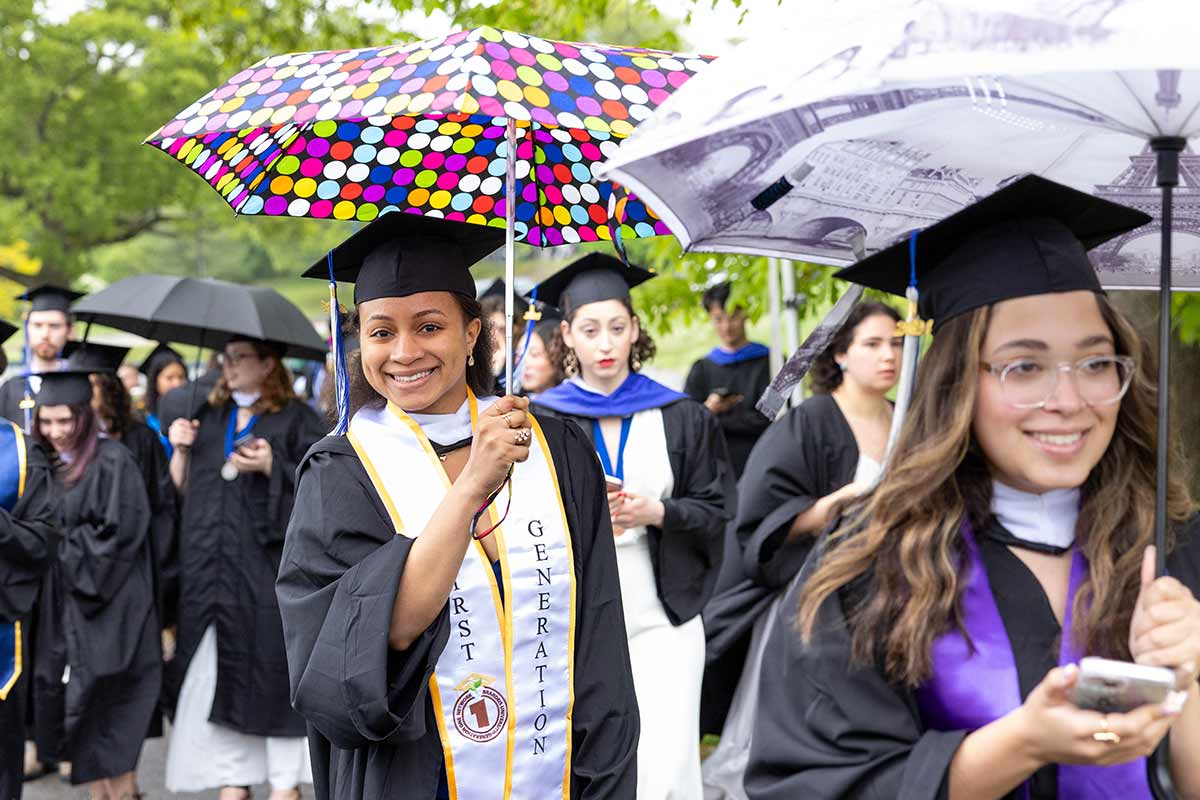A large group of graduates walk outside in the rain with umbrellas.