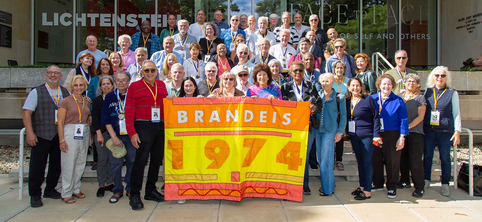 A group of alumni stand on the steps of the Rose Art Museum. The people in front are holding a Class of 1974 banner