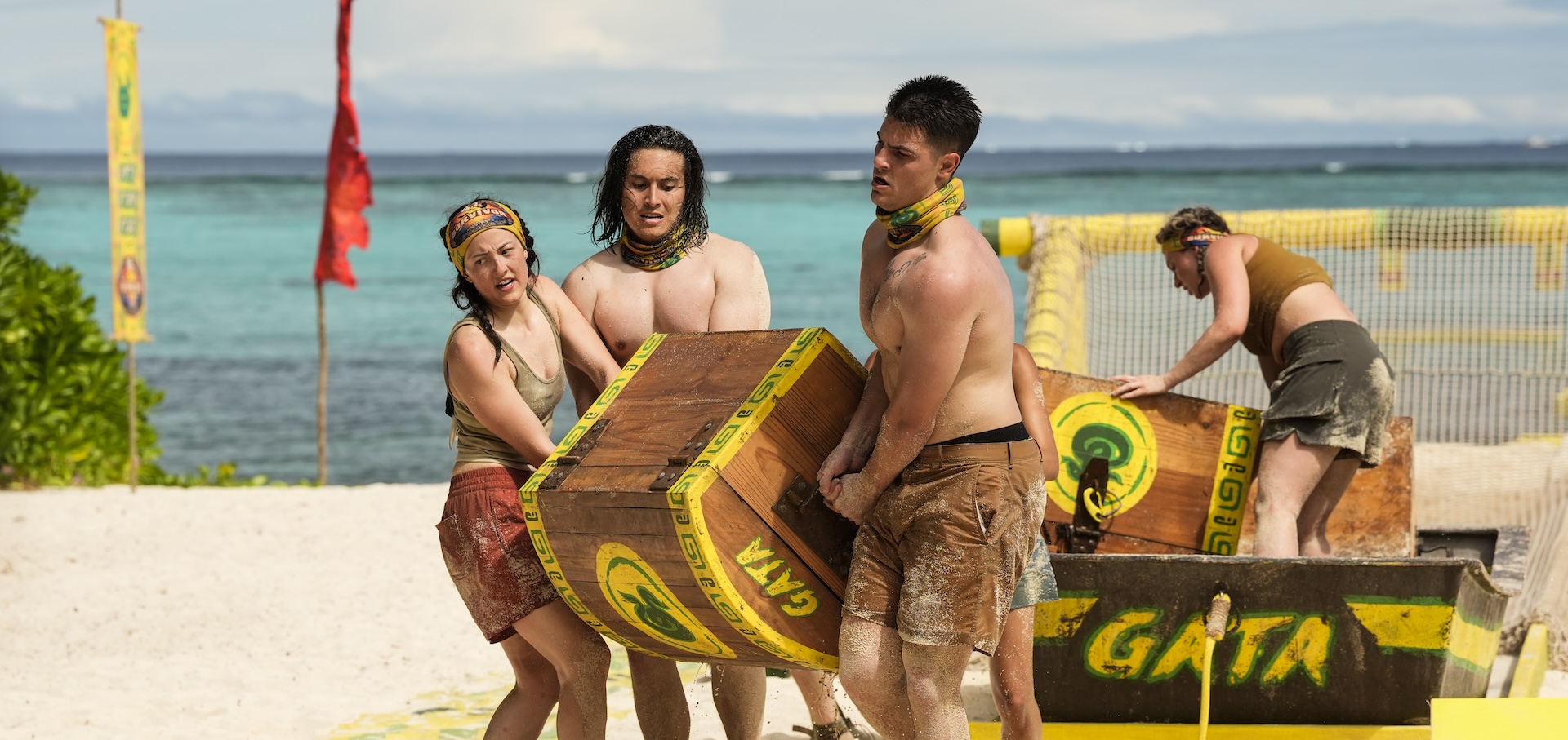 Four muscular contestants on the show "survivor" drag a wooden crate onto a beach.