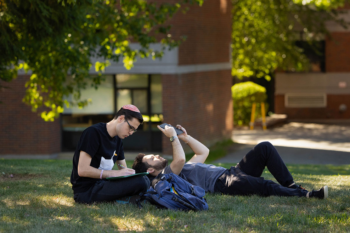 Two students sit outside on the grass on campus. One student is writing in a notebook and the other is reclining and looking at a tablet.