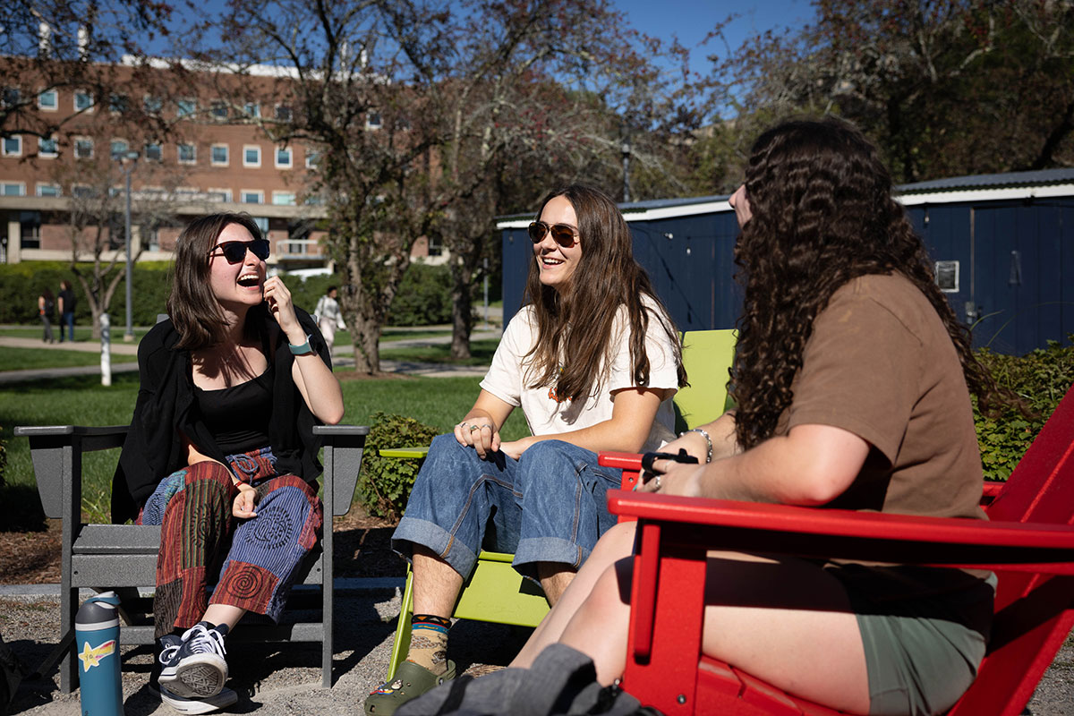 Three students sit in chairs outside on the Brandeis campus laughing.