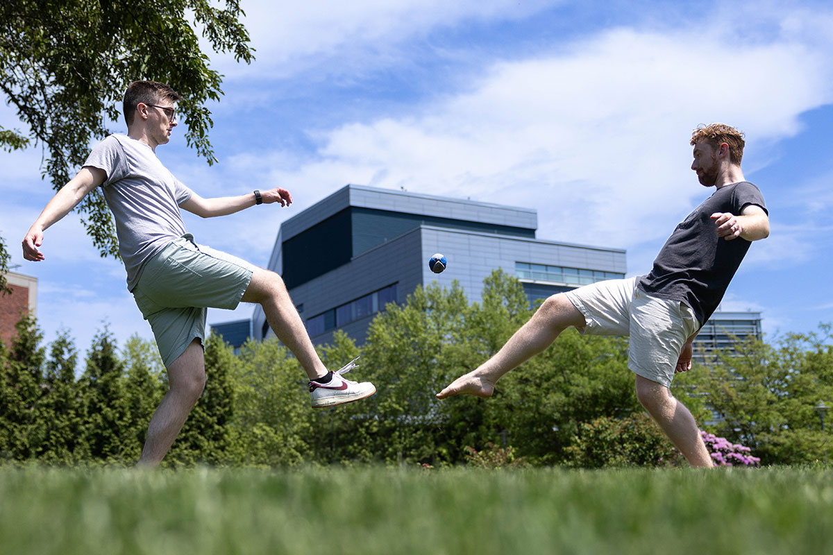 Two students play hacky sack outside on a sunny day.