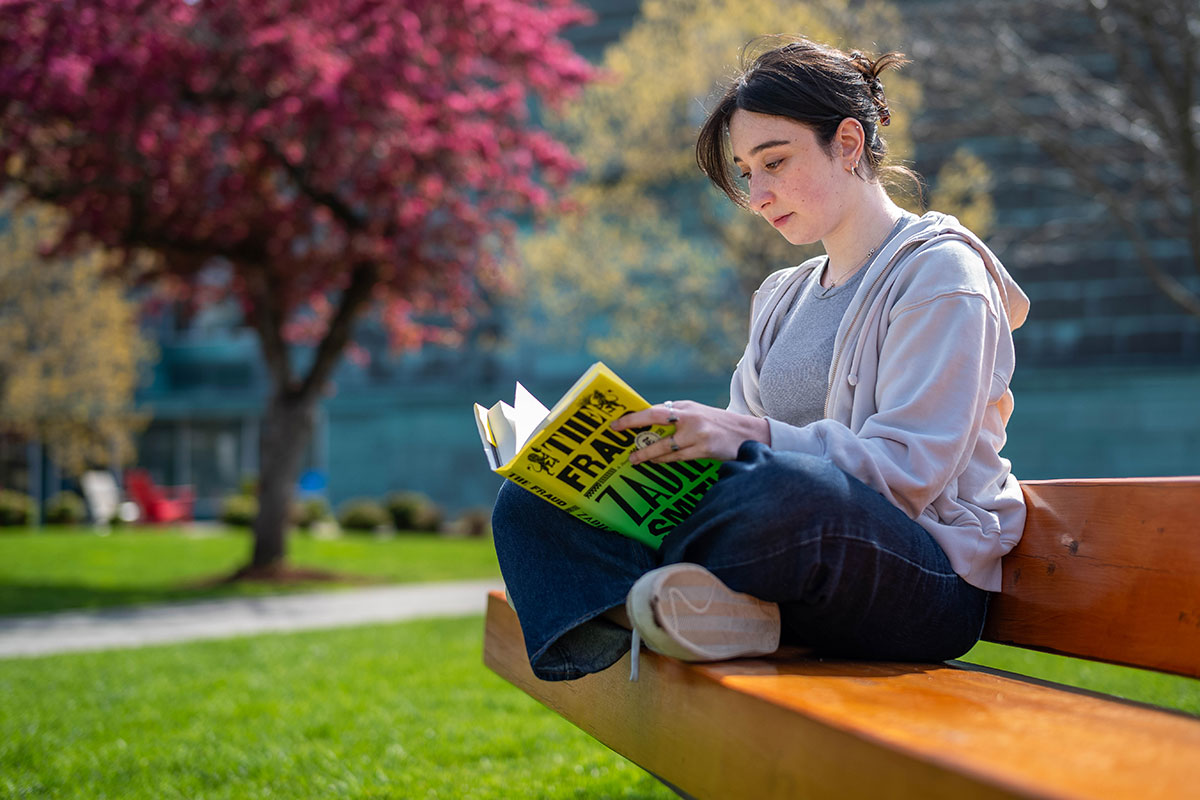 A student sits on a bench outside on a sunny spring day reading a book.