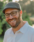 headshot of Uriah Kfir in a white shirt, hat and glasses, photographed outdoors with trees blurred in the background