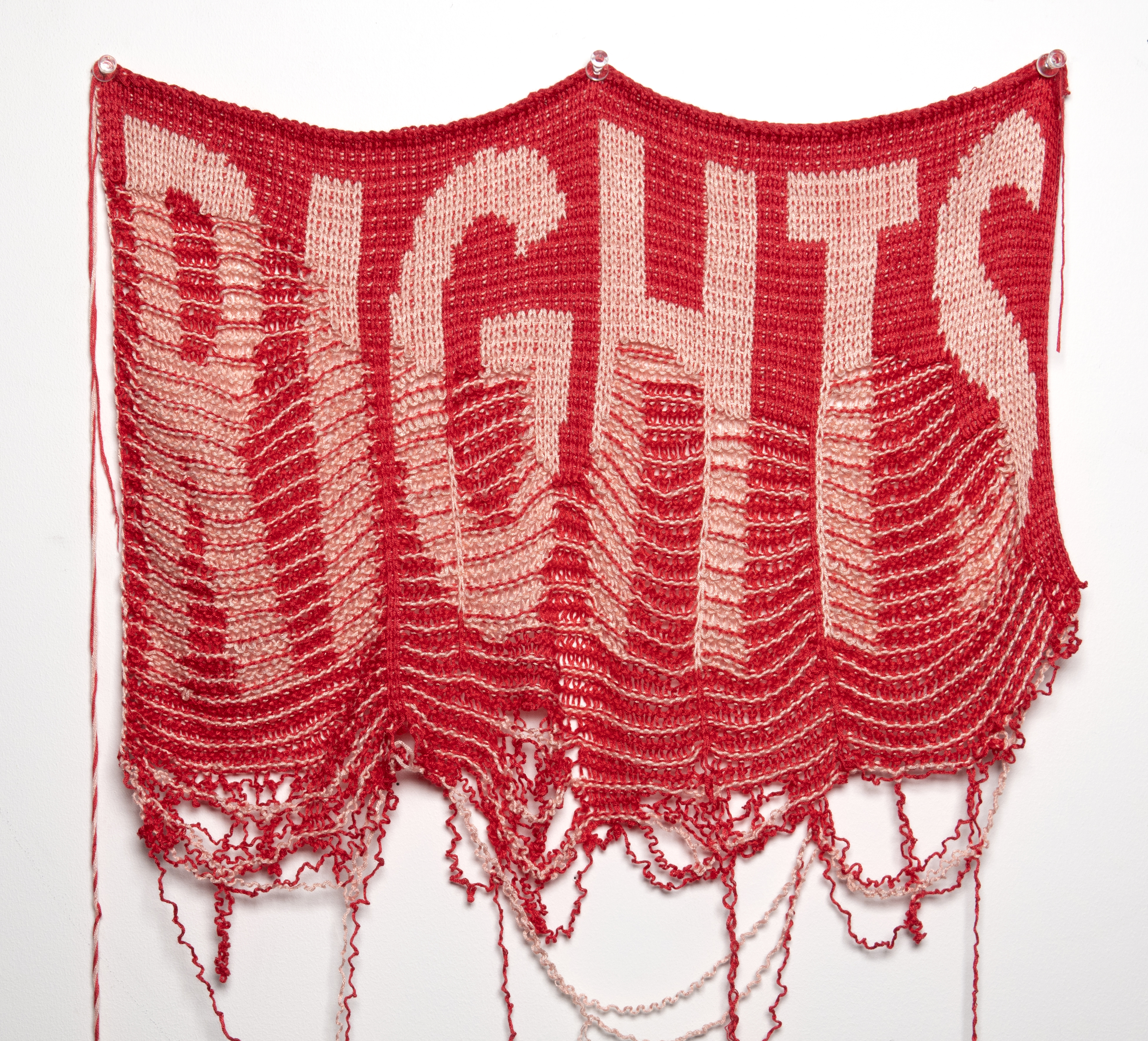 A video still of an unraveling red and pink knit cloth on which RIGHTS is inscribed 