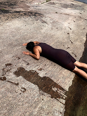 a figure in a black dress lays face down on the rock near the water. the sky is blue, there are wet body prints on the rock